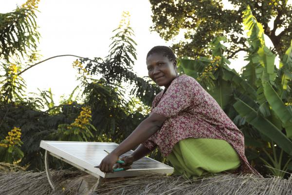 Eililoy Kamwendo smiles as she crouches on a thatched roof to fix a solar panel