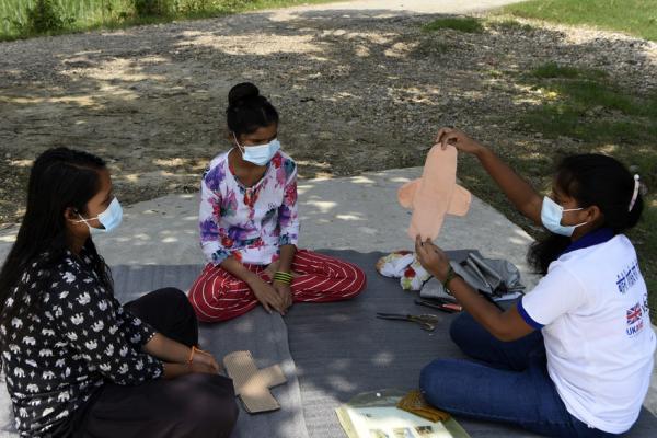 three woman sat on the floor outside. One is teaching how to make a reusable sanitary pad