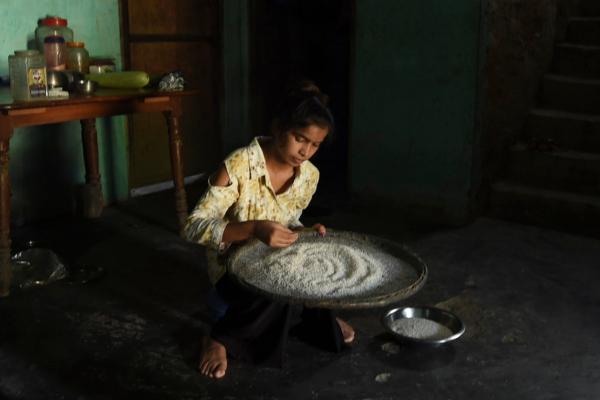 A young girl sits on a low stool in her family's home, examining a large dish of rice as part of dinner preparations