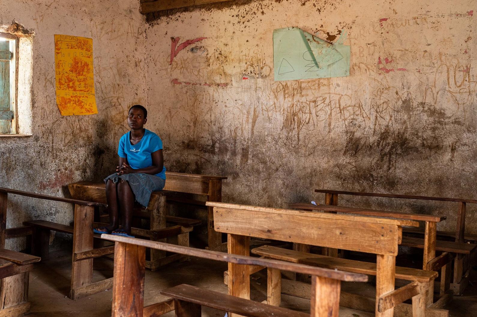 Schoolgirl Elizabeth sits on a wooden bench in a classroom, her hands clasped in front of her