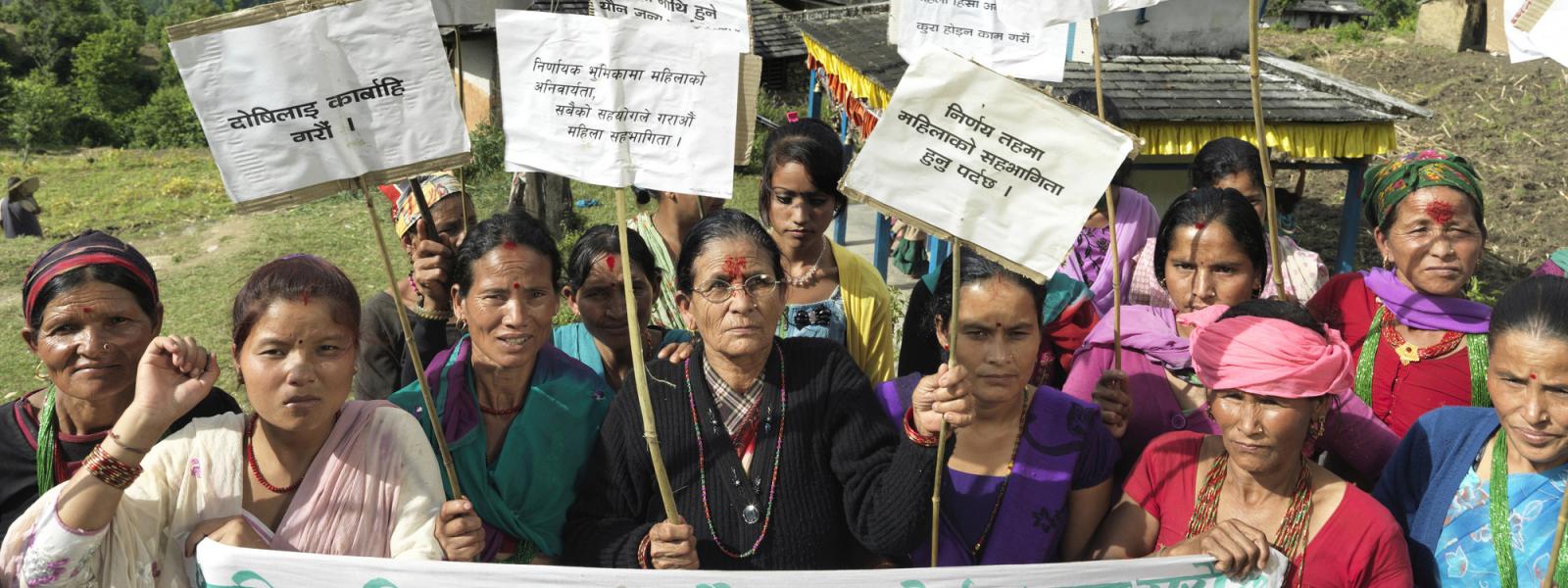 A group of Nepali women hold a banner and placards as they protest in front of their remote village, with hills in the background
