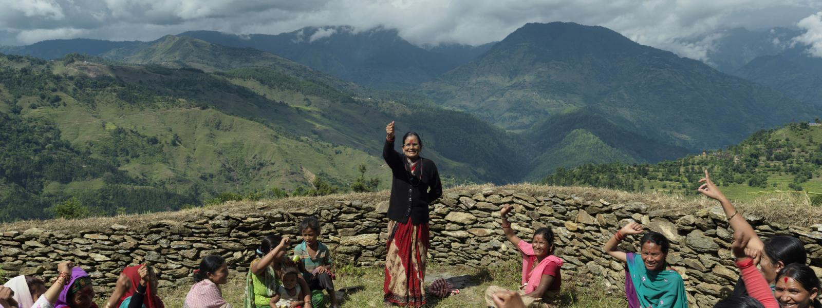 Women sit outside in a circle and raise their hands as one woman stands. In the background are steep rolling hills