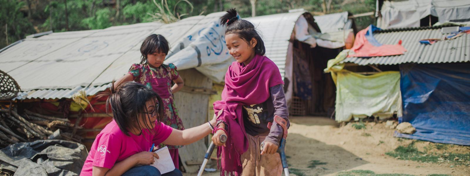 A volunteer crouches to help a young girl learn to use crutches, outside in Dhading camp for internally displaced peoples.
