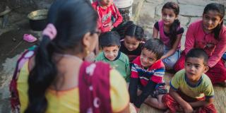 A group of young children sit outside on the round, listening to a local teacher talk about school and earthquake preparedness