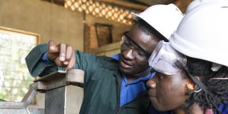 A student watches intently as her instructor demonstrates machinery in a welding and metal workshop