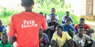 Peer educator Alfred Kunda stands in front of a group of seated young men, delivering an outdoor peer education session
