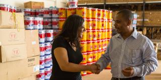 A man and woman shake hands in a warehouse, in front of stacks of drums of produce