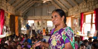 A female teacher speaks passionately at the front of a room full of people sitting down listening to her