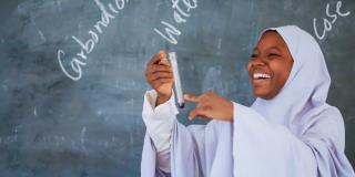 A female student stands in front of a blackboard and smiles as she examines a test tube