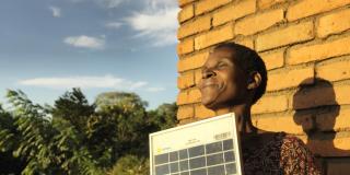 Solar Mama Dines smiles as she stands outside in the sun holding a solar panel