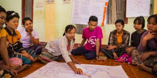 A group of people sit on the floor as Daw Hla Myint points out the map of village during a disaster risk management awareness session in Kyatkathone Village