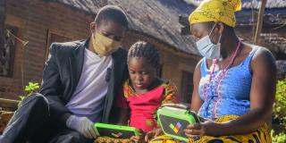 A young girl sits outside with her mother and father, who are both wearing face masks, using a tablet to learn