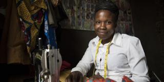 Tailor Macklyne Katsuiime sits at her sewing machine, with patterned fabric hanging next to her and a tape measure around her neck