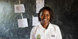 National volunteer Brown Niyonsaba stands smiling in front of the blackboard in a classroom at Umutara Deaf School in Rwanda.