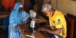 Two people sit inside at a table, wearing masks and using a sewing machine