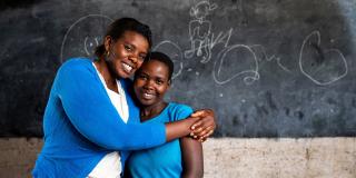 Mentor Regina stands with her arms round the shoulders of student Elizabeth, in front of a blackboard in the classroom