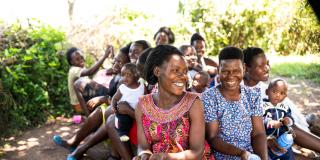 Teacher Christine and a group of students smile and laugh as they sit on the ground outside during breaktime