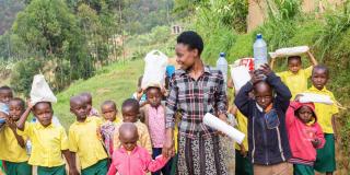 A group of young children carry teaching materials as they walk outside, led by their teacher