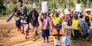Teacher Josephine Nyirampuhwe on her way to Boli Nursery School with some of her pupils, who are helping her to carry teaching materials A group of young children carry teaching materials as they walk outside, led by their teacher