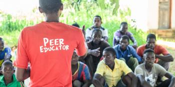 Peer educator Alfred Kunda stands in front of a group of seated young men, delivering an outdoor peer education session