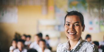 Primary school teacher Souy Kran smiles as she stands at the front of her classroom; behind her, rows of schoolchildren work at their desks.