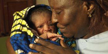 A midwife smiles gently as she holds a newborn babby swaddled in a colourful blanket