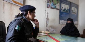 A female police officer sits across a desk from a civilian woman, in the police station