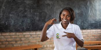 Volunteer Brown Niyonsaba smiles as she sits in front of a blackboard in the classrrom at Umutara School, signing 'VSO volunteer'