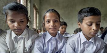 Two young girls and a young boy sit and read from their textbooks in a busy classroom