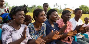 Women sit in a row and applaud as they listen to volunteer Juliana at a community meeting