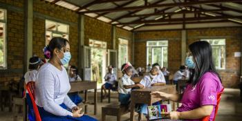 Community engagement volunteer Mi Hnin Hnin Wai speaks to a teacher as children in protective wear sit at their desks behind them