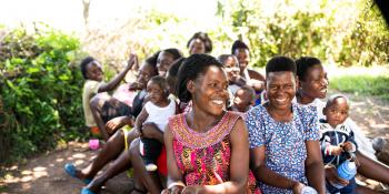 Teacher Christine and a group of students smile and laugh as they sit on the ground outside during breaktime