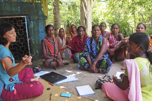 A group of women sit in a loose circle on the ground outside as a VSO volunteer sits and speaks with them