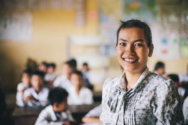 Primary school teacher Souy Kran smiles as she stands at the front of her classroom; behind her, rows of schoolchildren work at their desks.
