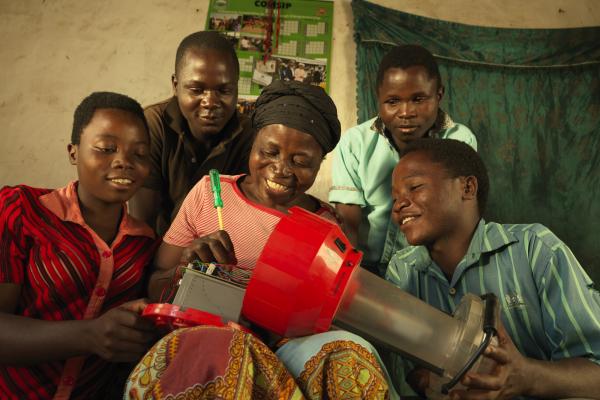A woman smiles as she uses a screwdriver to fix a large solar lantern, as a group of younger people gather round and watch.