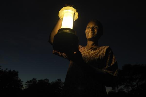 A woman's smiling face is illuminated by the solar lantern she holds up as she stands outside at nighttime