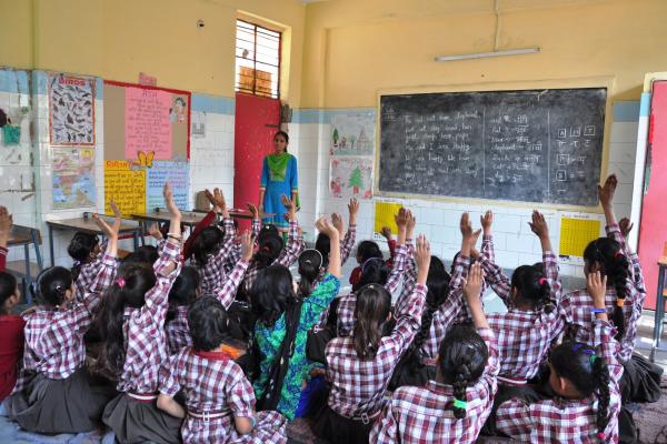Girls sit on the floor of their classroom and raise their hands; at the front, a young female teacher stands by the blackboard