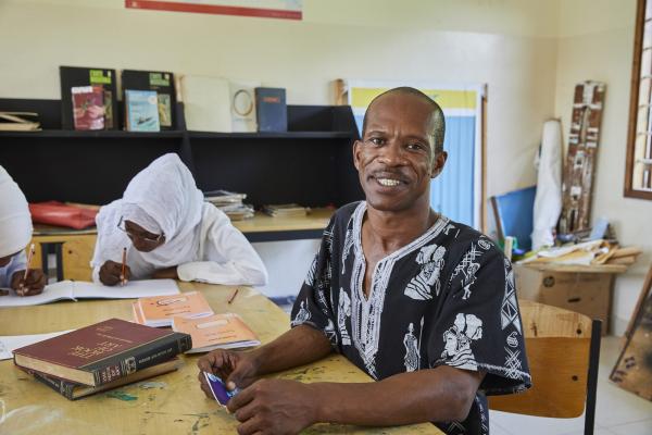 Art teacher Omar Kiwenge sits at a table in the studio, with students sketching behind him