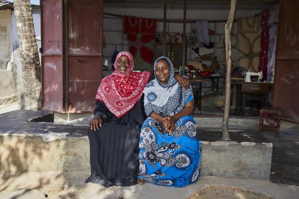 Two colourfully-dressed female tailors sit outside in front of their workshop