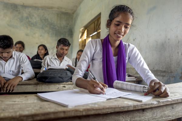 15-year-old Arti sits at her desk and works in her school book; behind her are more classmates studying