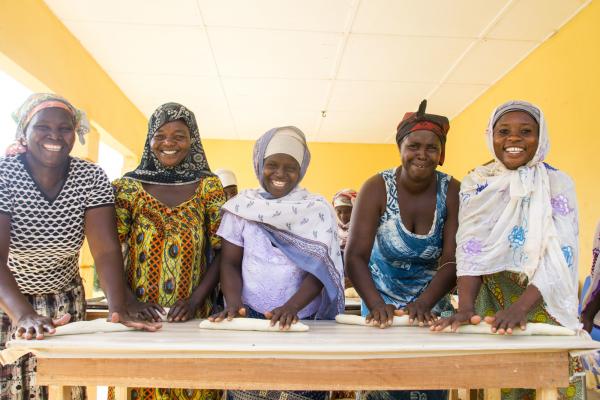 Five women smile as they roll out dough on a long table