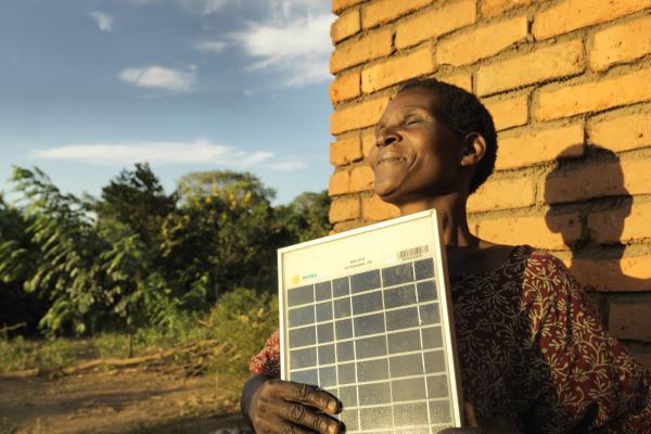 Solar Mama Dines smiles as she stands outside in the sun holding a solar panel