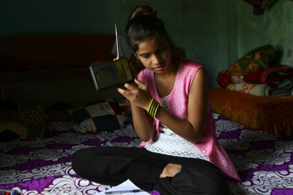 A young Nepali girl sits on her bed, holding up a portable radio to listen to an educational broadcast