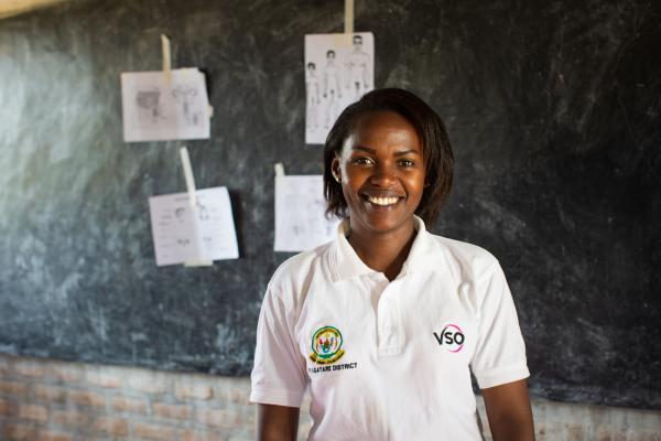 National volunteer Brown Niyonsaba stands smiling in front of the blackboard in a classroom at Umutara Deaf School in Rwanda.