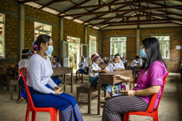 Community engagement volunteer Mi Hnin Hnin Wai speaks to a teacher as children in protective wear sit at their desks behind them