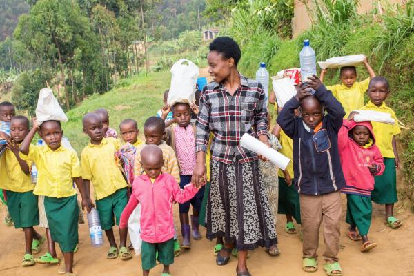A group of young children carry teaching materials as they walk outside, led by their teacher