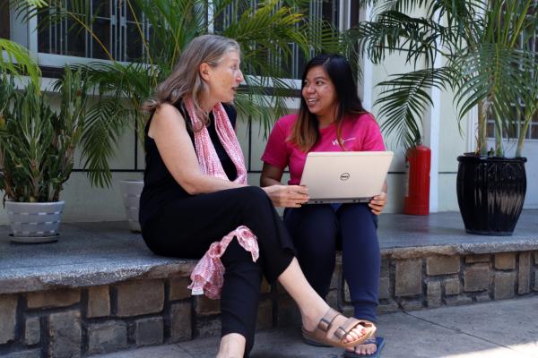 two women sat on a step outside a building talking to eachother with a laptop